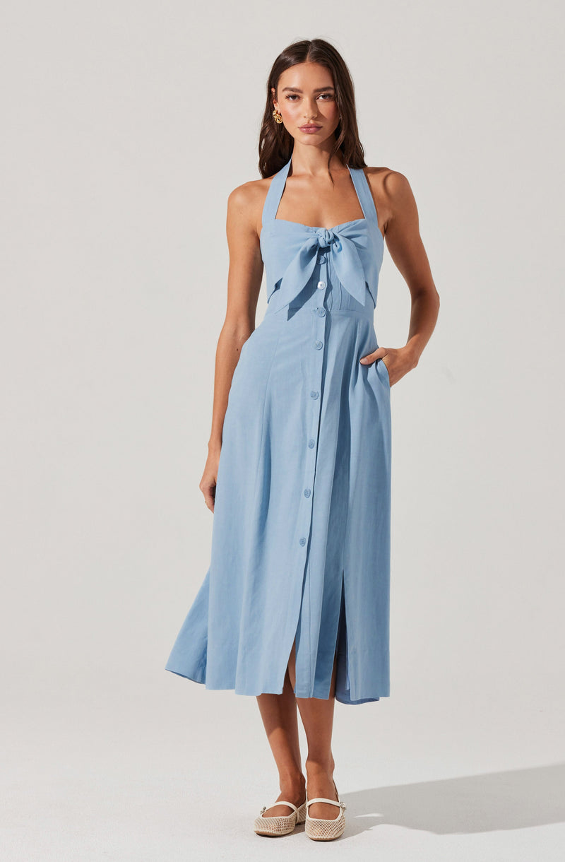 A woman is wearing a light blue sleeveless dress with a fitted bodice and a large bow at the front neckline. The dress features buttons running down the center and a slit at the front of the skirt. She has her right hand in her pocket and is wearing white woven shoes.