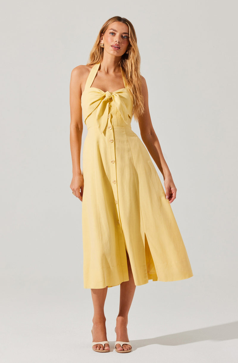 A woman is wearing a yellow sleeveless dress with a halter neckline and a large bow at the front. The dress features a button-down design running from the waist to the hem, with a slit in the front. She is also wearing open-toed beige high heel shoes.