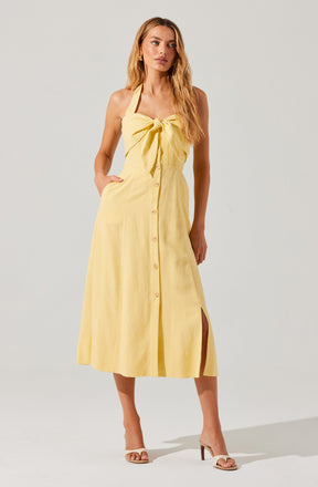 A woman is wearing a sleeveless, light yellow midi dress with a large bow detail at the front. The dress features a button-down front and a side slit. She is also wearing high-heeled sandals.