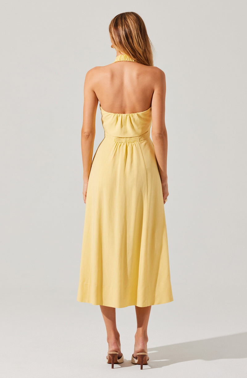 A woman is wearing a sleeveless, yellow halter-neck dress with an elastic waistband. The dress has a gathered bodice and a long, flowing skirt that reaches mid-calf. She is standing with her back to the camera, and her hair is styled in loose waves.