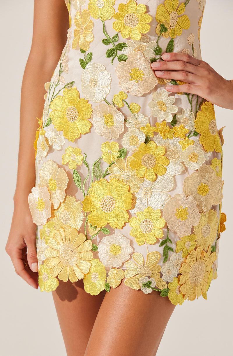 A close-up of a woman's dress featuring an intricate floral embroidery pattern with yellow, cream, and pastel pink flowers, along with green leaves. The dress is made of a sheer, light fabric with three-dimensional embroidered flowers covering the entire front. The woman's right hand is resting on her waist, while her left hand hangs down by her side.