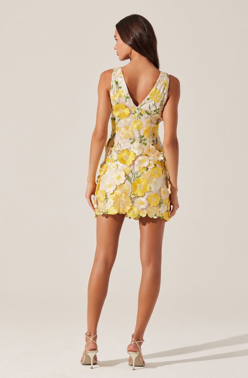 A woman is wearing a sleeveless dress with a V-neckline and a floral pattern featuring yellow, white, and green colors. The dress has a fitted waist and a short skirt that ends above the knees, with a scalloped hemline. She is also wearing beige high-heeled sandals with ankle straps.