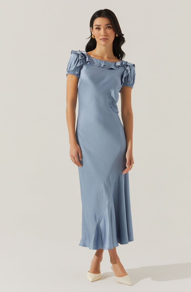 A woman is wearing a floor-length, light blue satin dress with puffed short sleeves that have ruffled details along the shoulders and neckline. The dress is fitted at the waist and has a slightly flared hem. She is also wearing off-white pointed-toe heels.