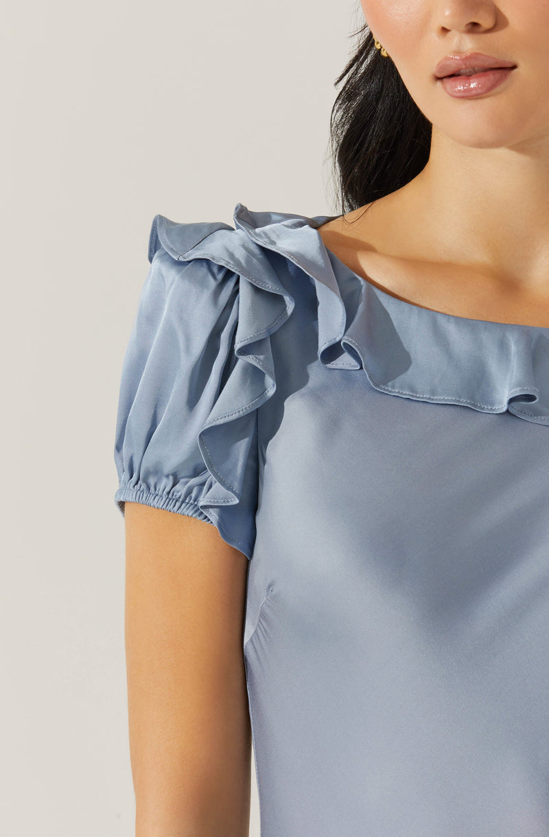 A woman is wearing a light blue satin top with ruffle details along the neckline and shoulder. The top has short, puffed sleeves with elasticized cuffs.