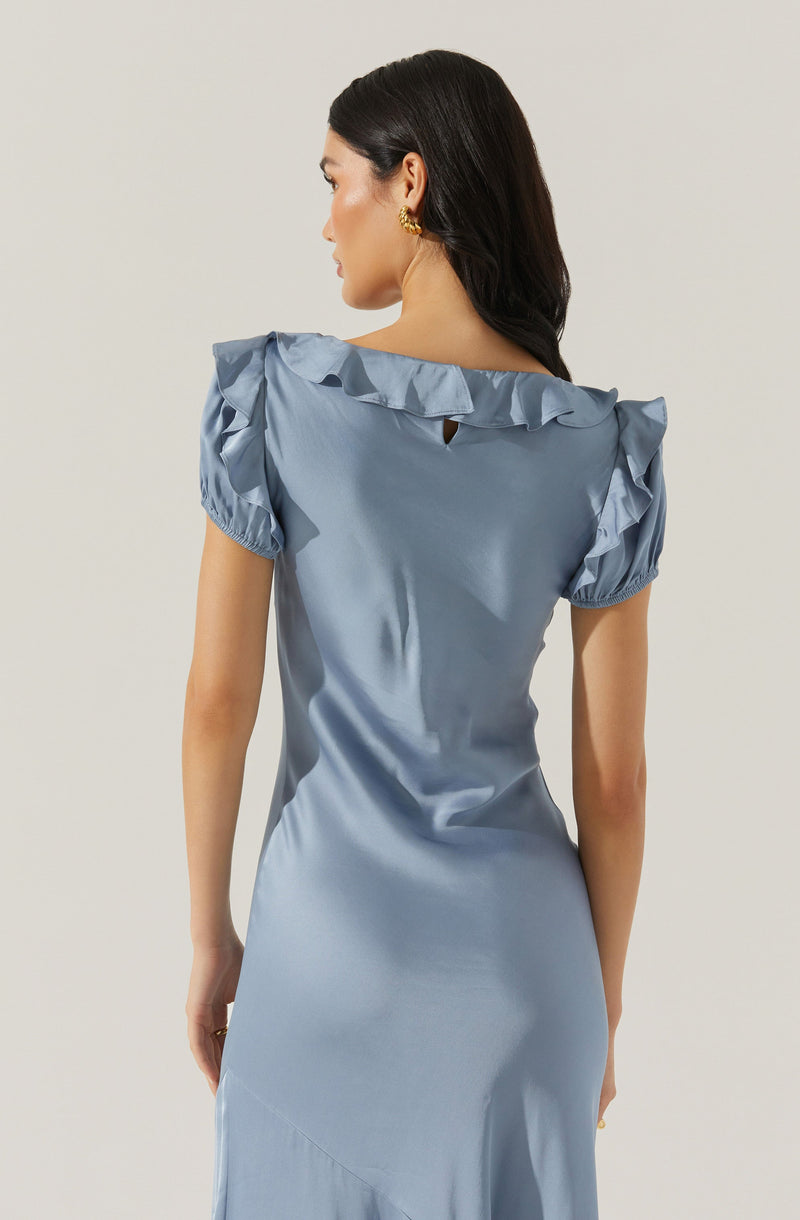 A woman is wearing a light blue satin dress with ruffled cap sleeves. The dress has a slight sheen and features a small keyhole opening at the back neckline. She has dark, wavy hair and is accessorized with gold earrings.
