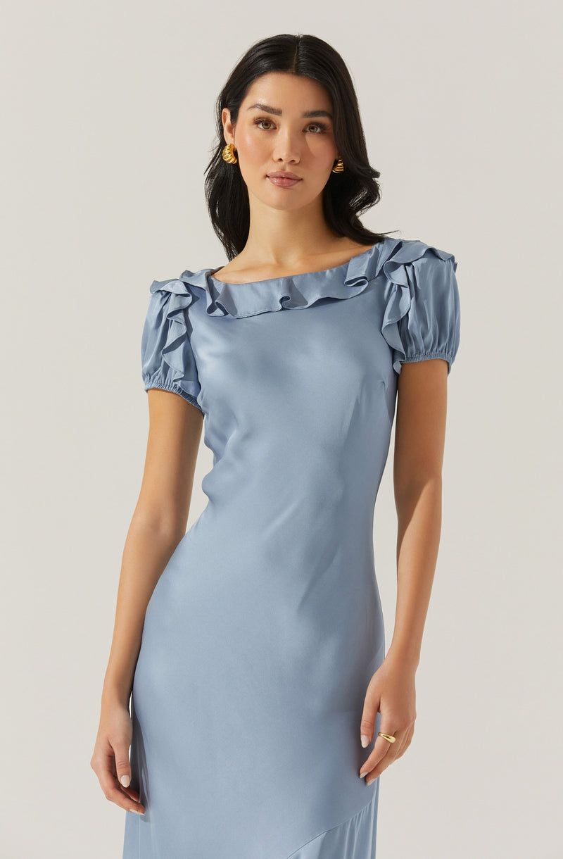 A woman is wearing a light blue satin dress with ruffled details along the neckline and puffed short sleeves. The dress has a smooth, shiny texture and a fitted silhouette. She accessorizes with gold earrings and a ring.