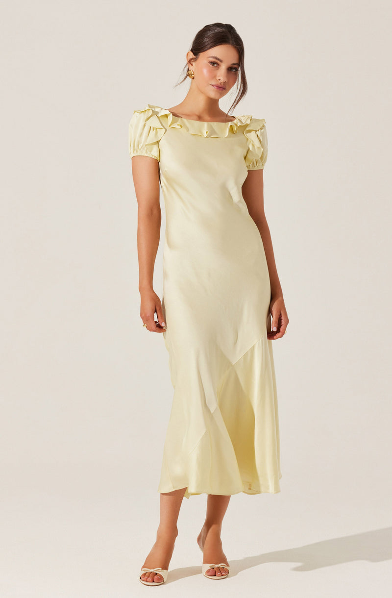 A woman is wearing a light yellow, full-length dress with short, puffy sleeves that have ruffle details. The dress has a round neckline with ruffled embellishments along the collar. She is also wearing open-toed, high-heeled sandals in a light color.