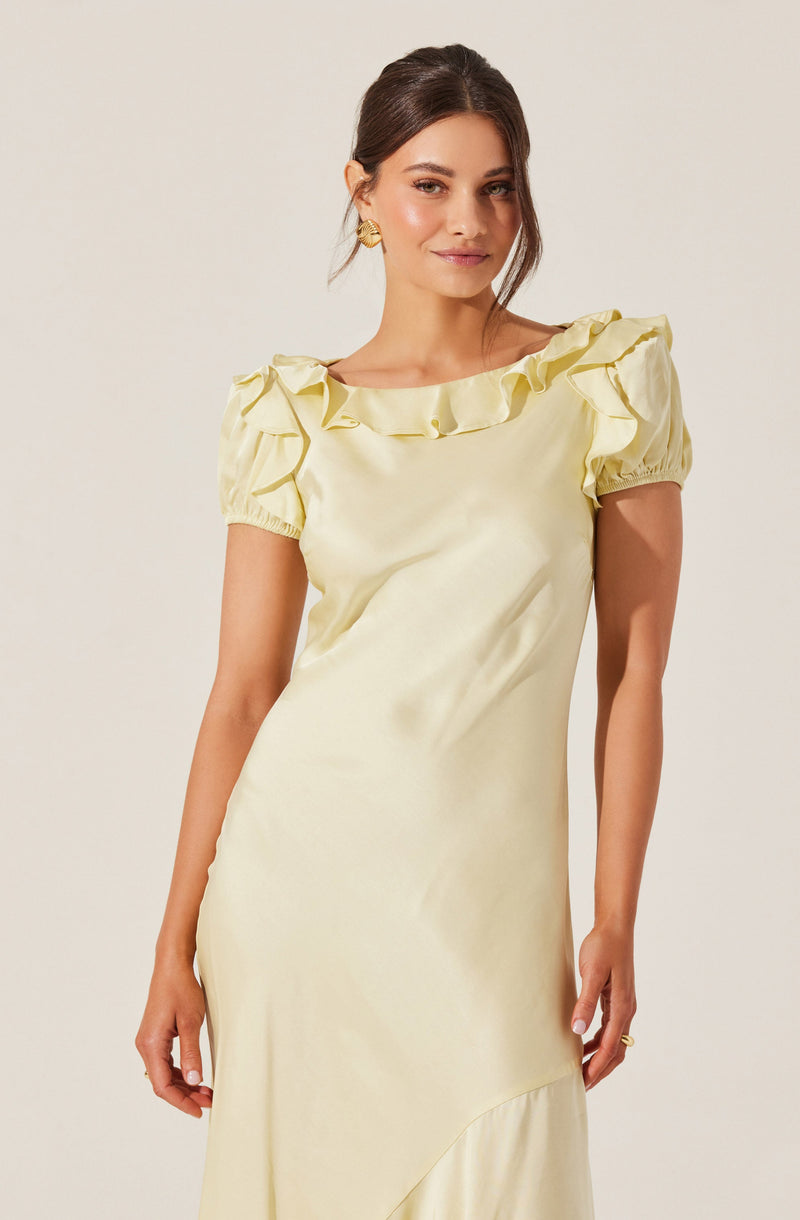 A woman is wearing an elegant cream-colored satin dress with a round neckline and short, puffed sleeves. The dress features ruffled details along the neckline. She has dark brown hair styled in a loose updo, and is accessorized with gold earrings.