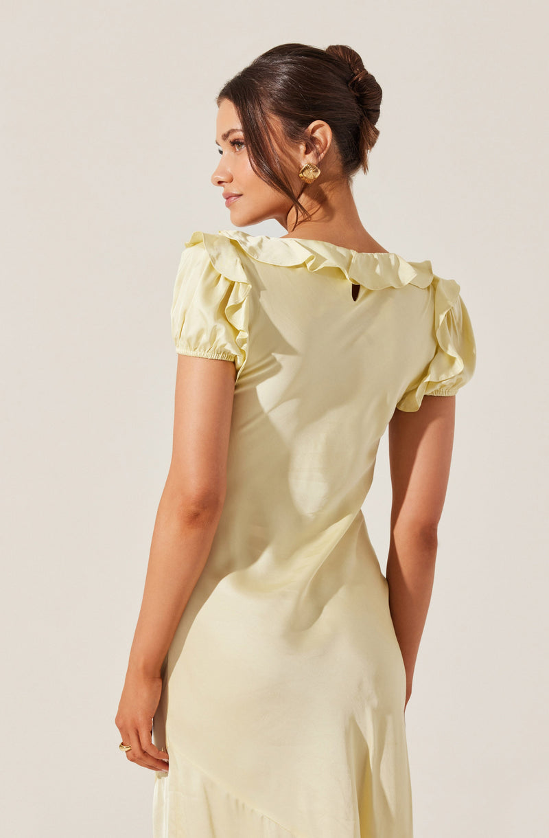 A woman is wearing a light yellow satin dress with puffed short sleeves that have ruffled edges along the shoulder and sleeve hem. The dress has a smooth, shiny texture and a fitted silhouette. She has her hair styled in an elegant updo and is accessorized with gold earrings and a ring.