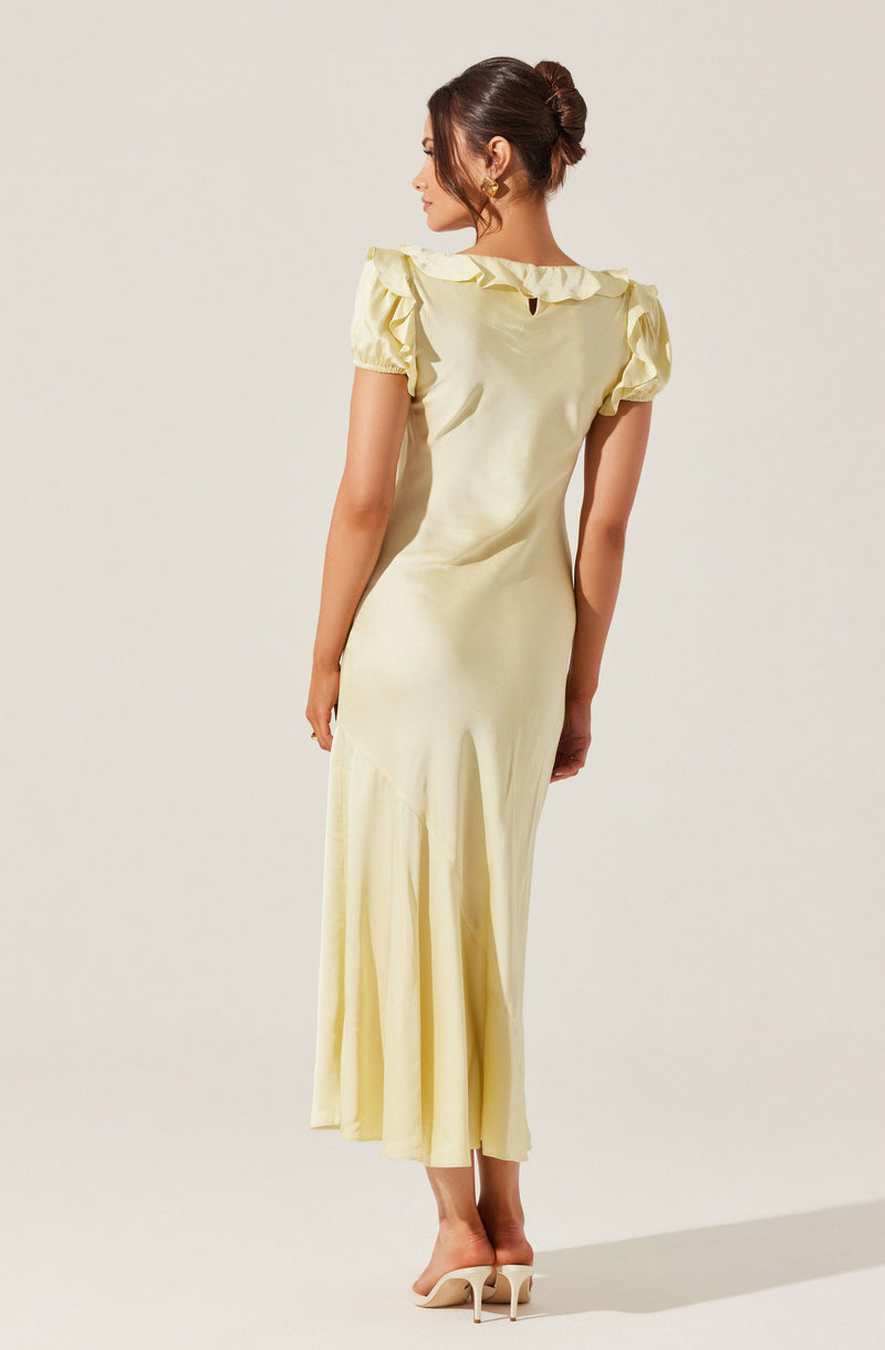 A woman is wearing a long, pale yellow dress with short puffed sleeves and ruffled detailing along the neckline. The dress has a fitted bodice and a flowing skirt that reaches the floor. She is also wearing nude high-heeled shoes.