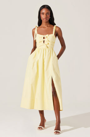 A woman is wearing a sleeveless, light yellow midi dress with shoulder tie straps. The bodice features a ruched design with multiple horizontal gathers and a front tie detail. The skirt has a high slit on the left side, revealing part of her leg. She is also wearing white open-toe high heels.