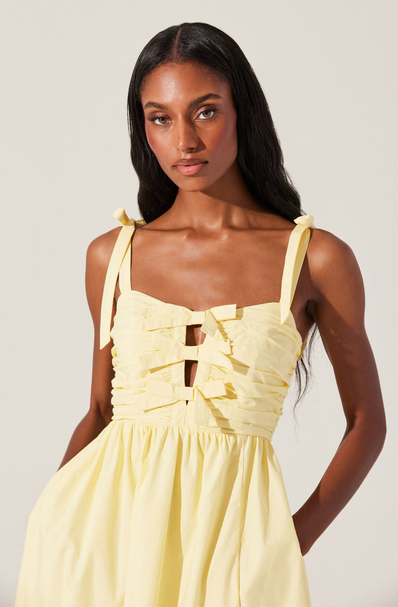 A woman is wearing a sleeveless, light yellow dress with shoulder straps tied into bows. The dress features a ruffled, ruched bodice with cut-out details in the center. The skirt is gathered and flows down from the waist.