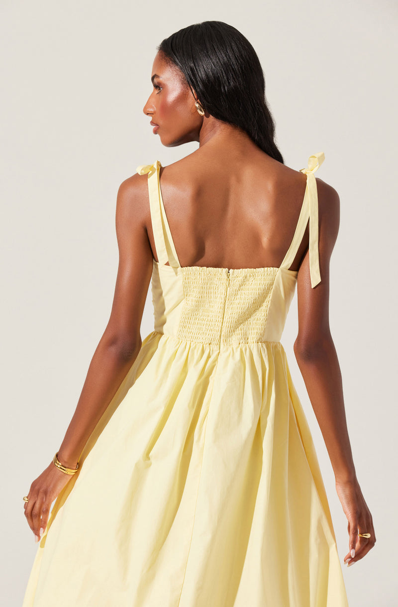 A woman is wearing a light yellow sundress with thin shoulder straps tied into small bows on her shoulders. The dress features a gathered bodice with a smocked panel at the back and a full, flowing skirt.