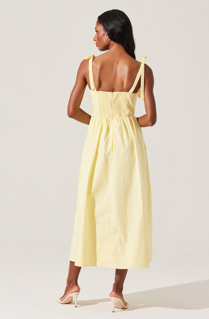 A woman is wearing a sleeveless, yellow sundress with shoulder ties and smocking detail on the bodice. The dress has a full, ankle-length skirt with pleats. She pairs the dress with white high-heeled sandals.