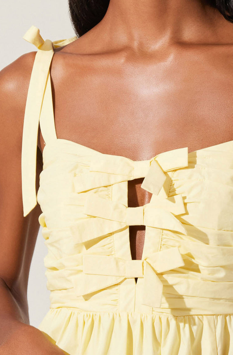 A woman is wearing a sleeveless, light yellow dress with shoulder straps tied into bows. The dress features vertical fabric bows on the front, creating a decorative and textured look. The fabric appears soft and ruched around the midsection, with a gathered skirt at the bottom.