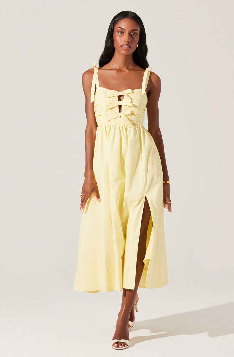 A woman is wearing a light yellow, sleeveless dress with shoulder straps tied into bows. The top part of the dress features ruffled detailing and cut-out accents. The skirt is long, flowing, and has a high slit on one side. She is also wearing white high-heeled sandals and has a gold bracelet on her right wrist.