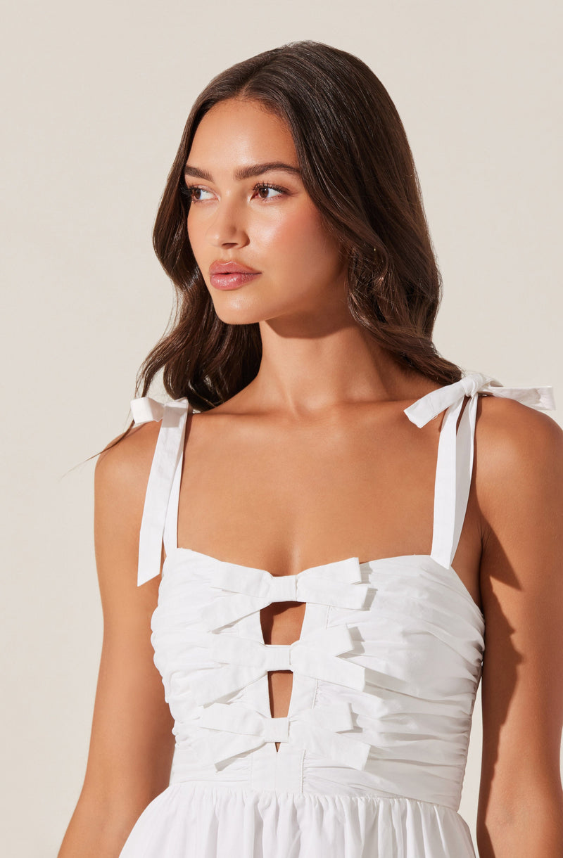 A young woman is wearing a sleeveless white dress with shoulder straps tied into bows. The bodice features a ruched design with horizontal gathers and three rectangular cutouts aligned vertically down the center, connected by horizontal fabric strips.