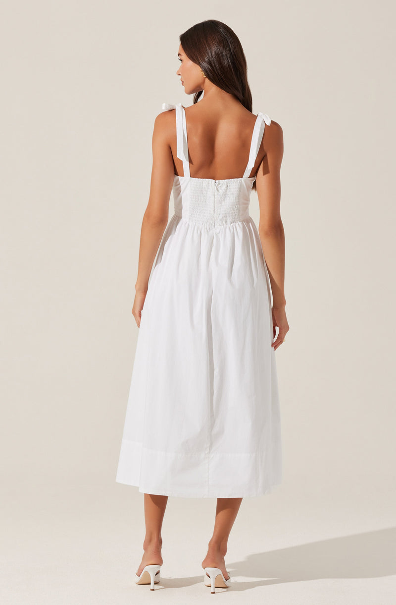 A woman is wearing a sleeveless, white dress with shoulder ties that form bows. The dress has a fitted, smocked bodice and a full, midi-length skirt. She pairs the dress with white heeled sandals.