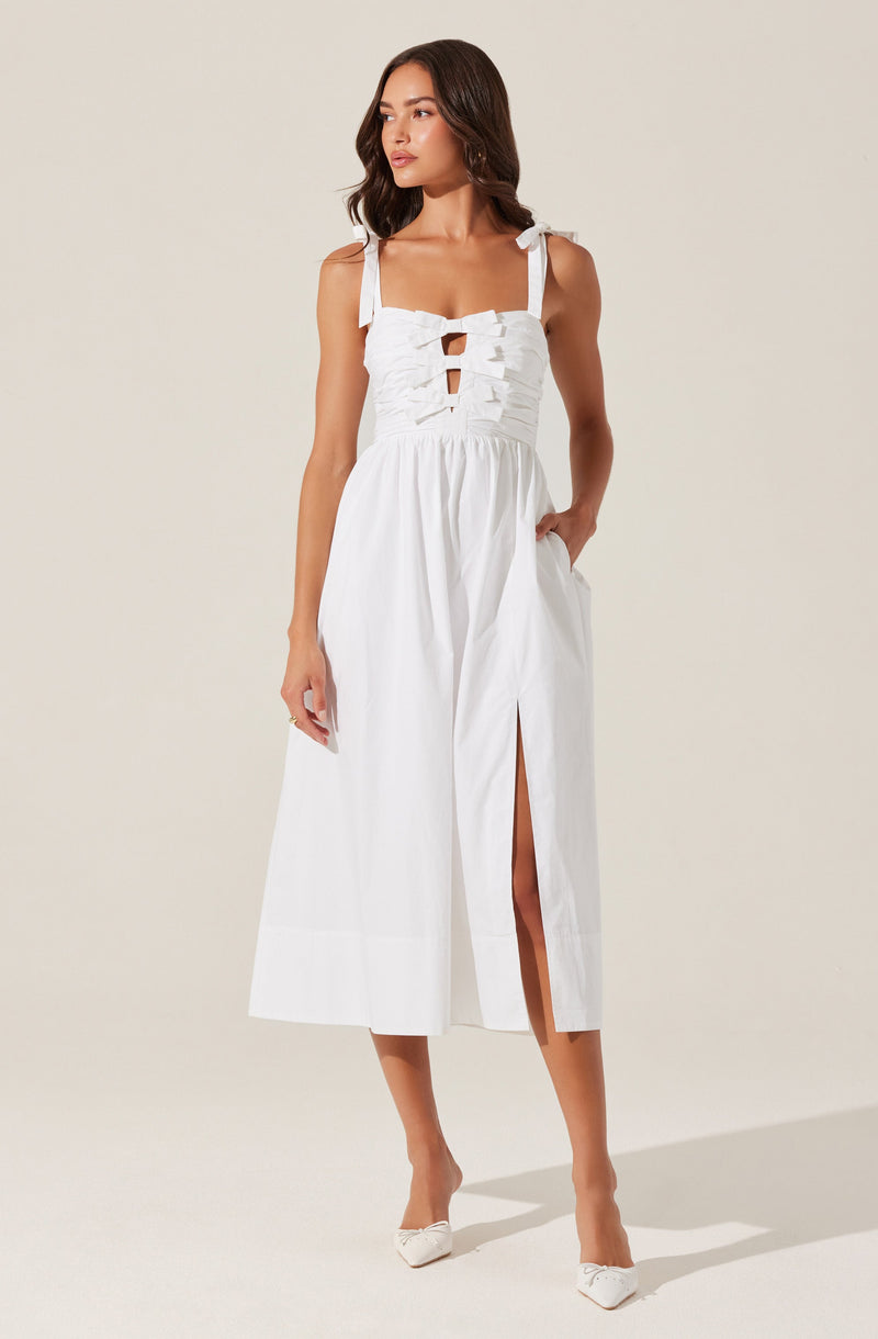 A woman is wearing a white sleeveless dress with shoulder straps tied into bows. The bodice features a ruched design with cut-out details in the front. The dress has a high waistline, a long skirt with a front slit, and pockets on each side. She is also wearing white pointed-toe heels.