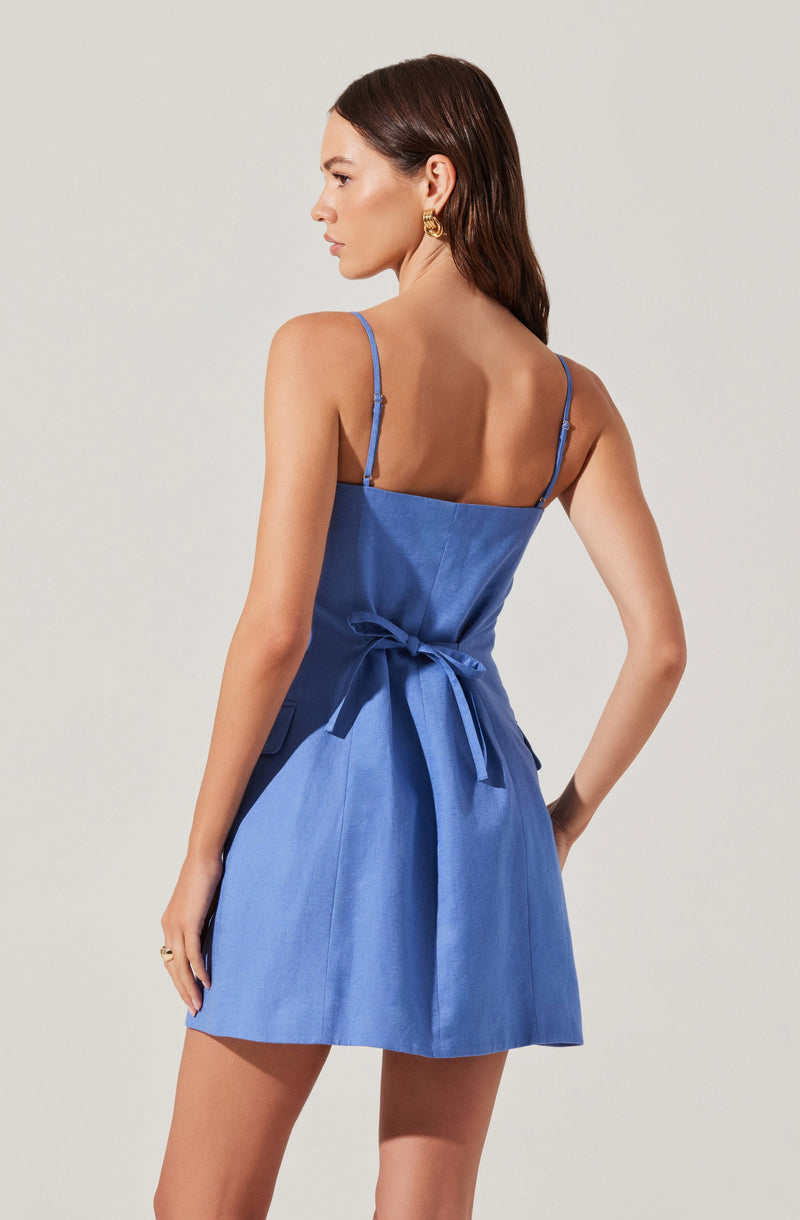 A woman is wearing a blue sleeveless dress with thin spaghetti straps. The dress features a fitted waist with a fabric belt tied in a bow at the back and has a slightly flared skirt. The fabric appears to be a smooth, shiny material.