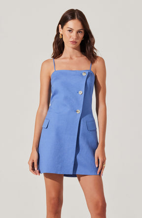 A woman is wearing a sleeveless, light blue dress with thin spaghetti straps. The dress features an asymmetrical front with three visible buttons running diagonally from the top left to the bottom right. It has two small, flap pockets on the front, positioned at the hips. The fabric appears textured, and the dress has a tailored fit that falls above the knees.
