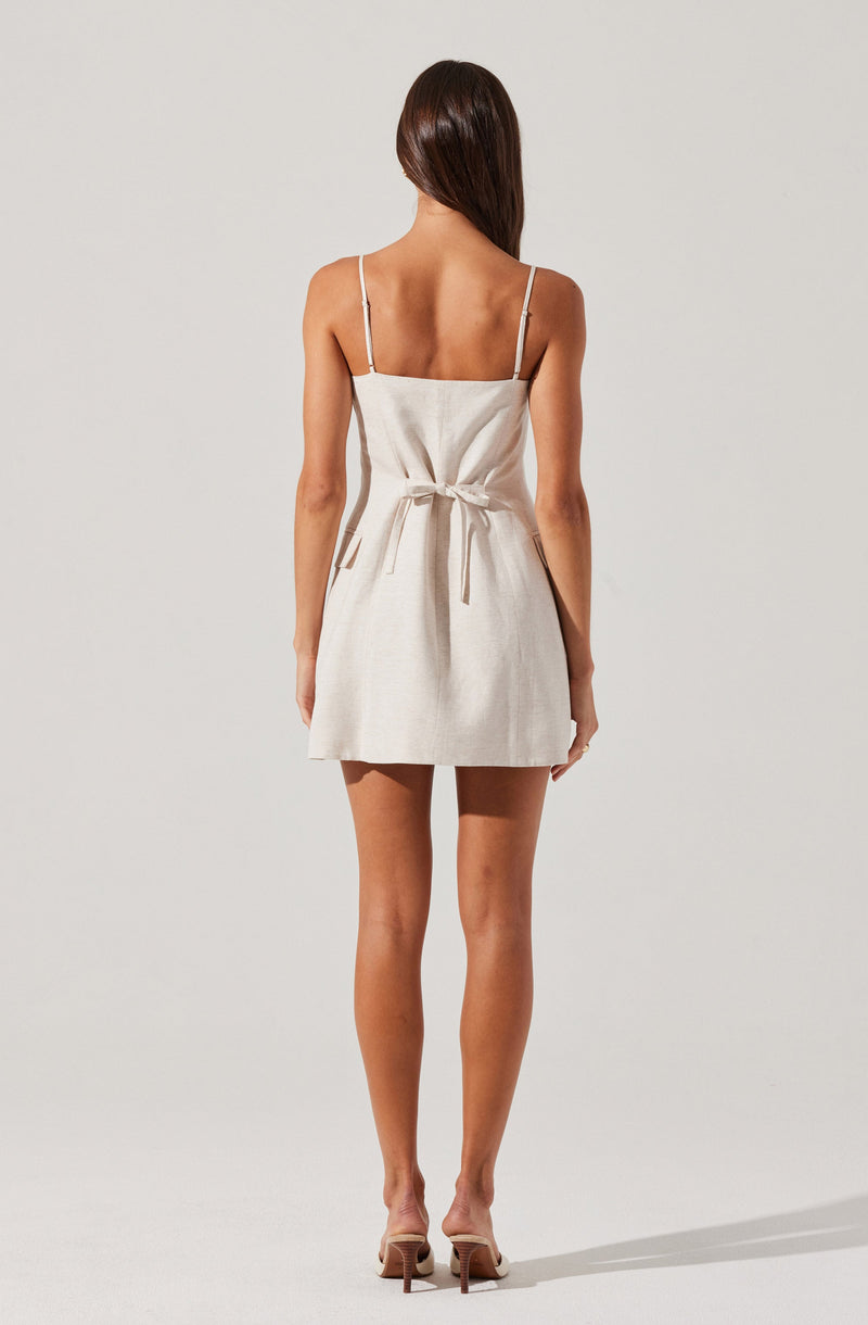 A woman is wearing a sleeveless dress with thin spaghetti straps. The dress is light-colored, possibly beige or off-white, and has a fabric belt tied at the waist. The hemline falls above the knees. She is also wearing nude-colored high-heeled shoes.