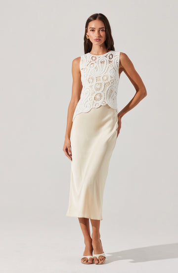 A woman is wearing a sleeveless, white lace top with intricate circular and floral patterns and a scalloped hem. She pairs the top with a long, smooth, satin-like cream-colored skirt that reaches her ankles. She completes the outfit with open-toed white heels.