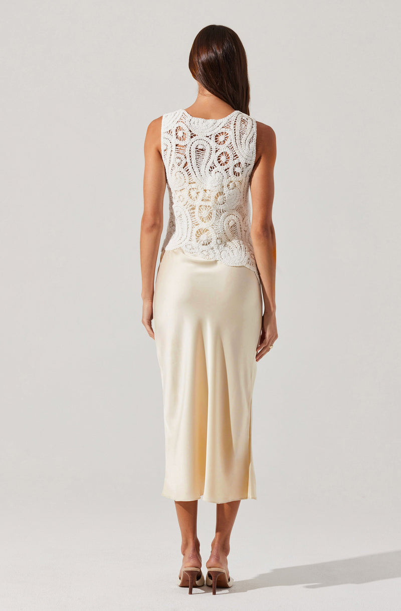 A woman is wearing a sleeveless white crochet top with intricate patterns and a round neckline. She pairs it with high-waisted, wide-leg, satin-like cream-colored pants that fall to her mid-calf. She is also wearing beige high-heeled sandals.