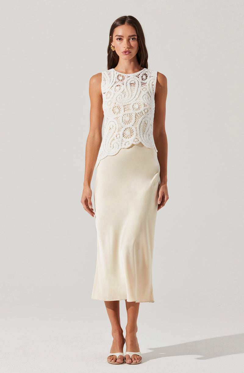 A woman is wearing a sleeveless, white lace top with intricate circular and floral patterns, paired with a long, smooth, ivory satin skirt that reaches her mid-ankles. She is also wearing nude-colored strappy heels and gold earrings.