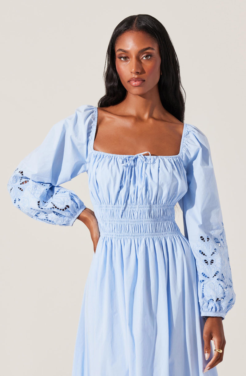 A woman is wearing a light blue dress with a square neckline and long, puffed sleeves. The dress features embroidery and cut-out details on the sleeves, with an elasticized waist creating a gathered effect.