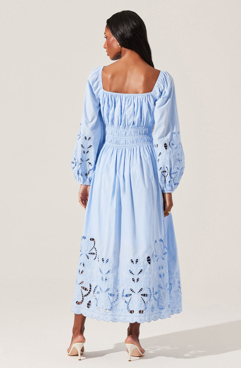 A woman is wearing a light blue, long-sleeved, ankle-length dress with puffed sleeves that feature eyelet embroidery. The dress has a gathered, elasticized waist and a square neckline, with intricate cutout lace details along the hem and sleeves. She is also wearing white high-heeled shoes.