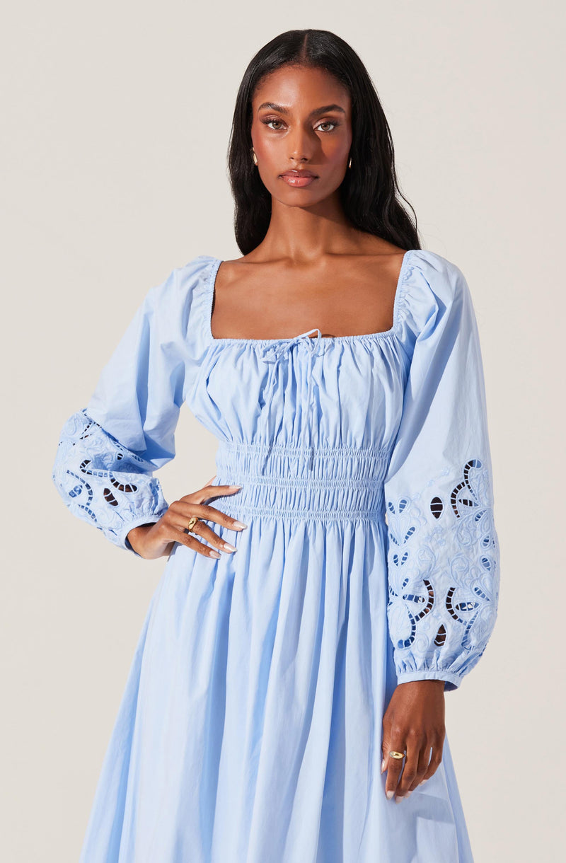 A woman is wearing a light blue dress with a square neckline and puffed long sleeves. The sleeves feature intricate eyelet embroidery with cut-out patterns near the cuffs. The bodice of the dress is gathered with elastic shirring, creating a fitted look at the waist, and the skirt is flowy with vertical pleats.