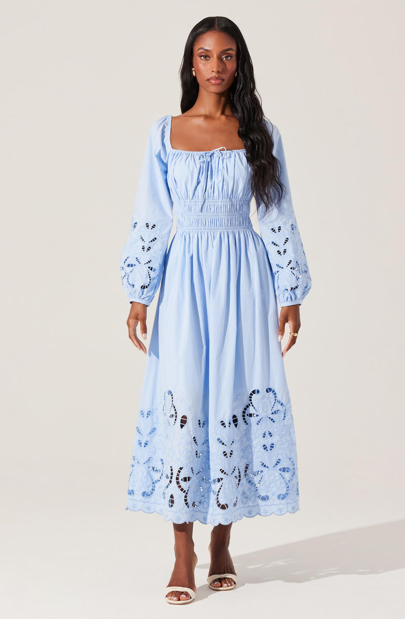 A woman is wearing a light blue, long-sleeved dress with intricate cutout embroidery. The dress has puffed sleeves with embroidered details, a fitted, gathered bodice, and a flowing skirt that reaches her ankles. The skirt and sleeves feature decorative cutouts with floral and geometric patterns, adding texture and visual interest. She pairs the dress with nude high-heeled sandals.