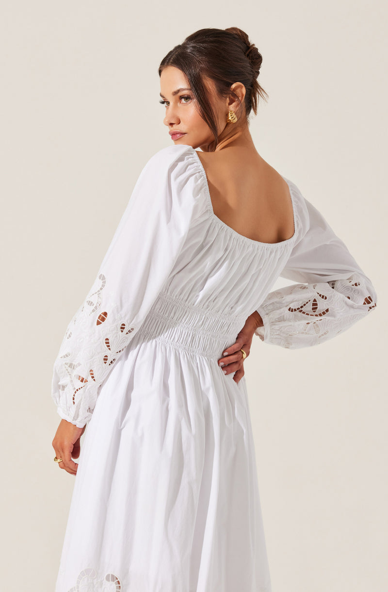 A woman is wearing a long white dress with a square neckline. The dress features puffed long sleeves with embroidered cutout details. The waist of the dress is cinched with gathered, elasticized smocking, and the fabric appears lightweight and airy. She has brunette hair styled in an elegant updo and is wearing gold earrings.