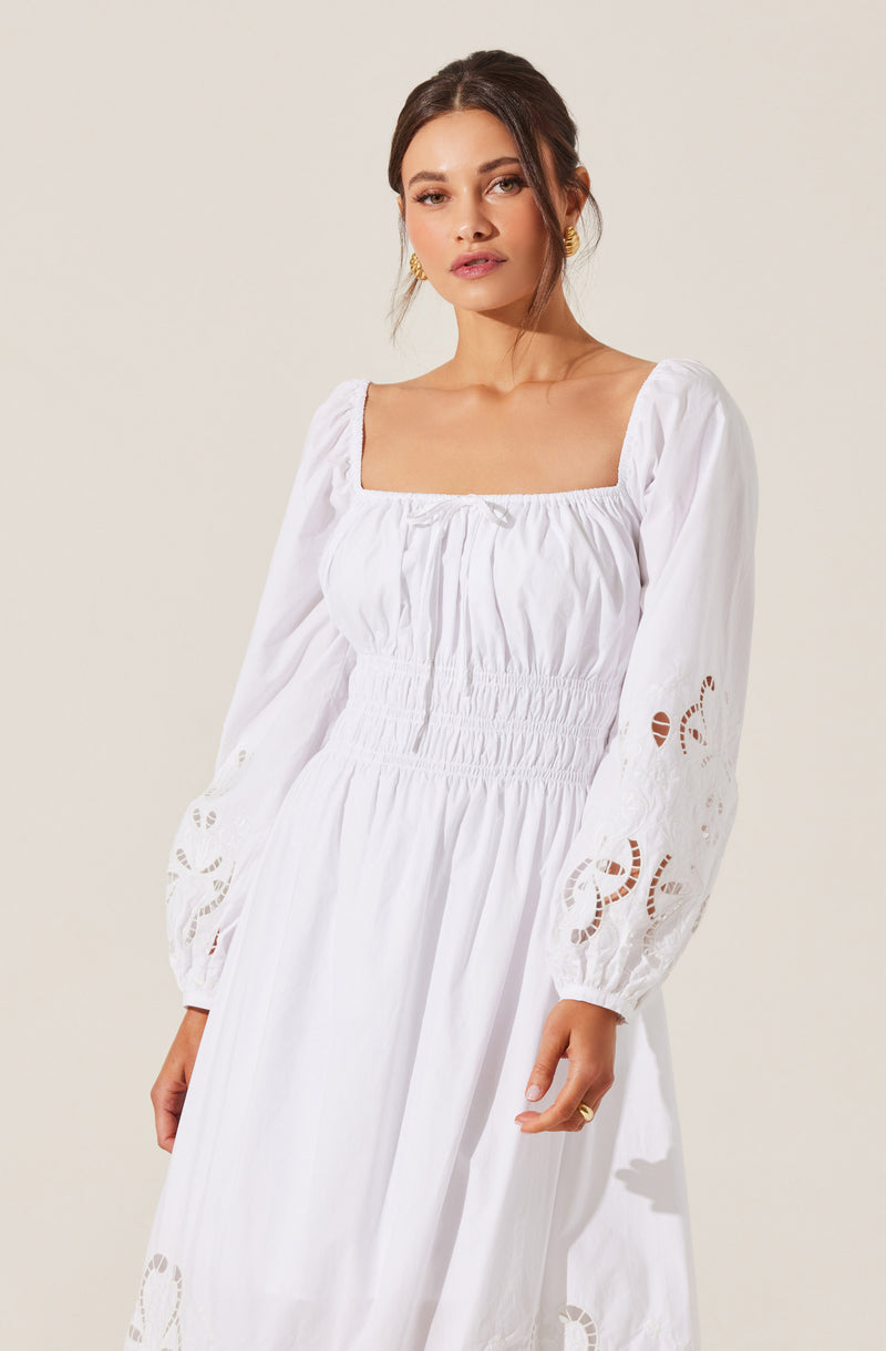 A woman is wearing a long white dress with a square neckline, puffed long sleeves with intricate embroidery cut-outs near the cuffs, and a gathered waistband with shirring detail. The dress appears to be made of a lightweight, flowy fabric.