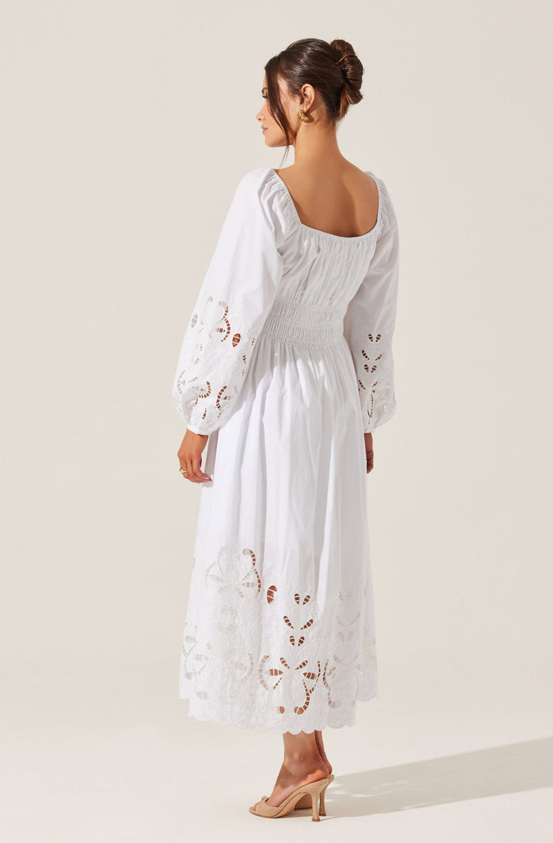 A woman is wearing a white, long-sleeved dress with intricate cut-out embroidery details on the sleeves and hemline. The dress has a fitted, ruched bodice with a square neckline and a flowing skirt that falls to her ankles. She is also wearing beige high-heeled shoes.