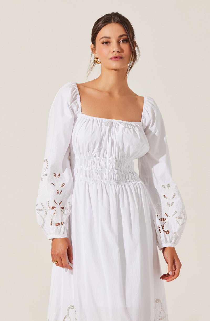 A woman is wearing a white, long-sleeved dress with a square neckline. The dress features embroidered cut-out designs on the sleeves and skirt, and has a smocked waist that creates a fitted silhouette.