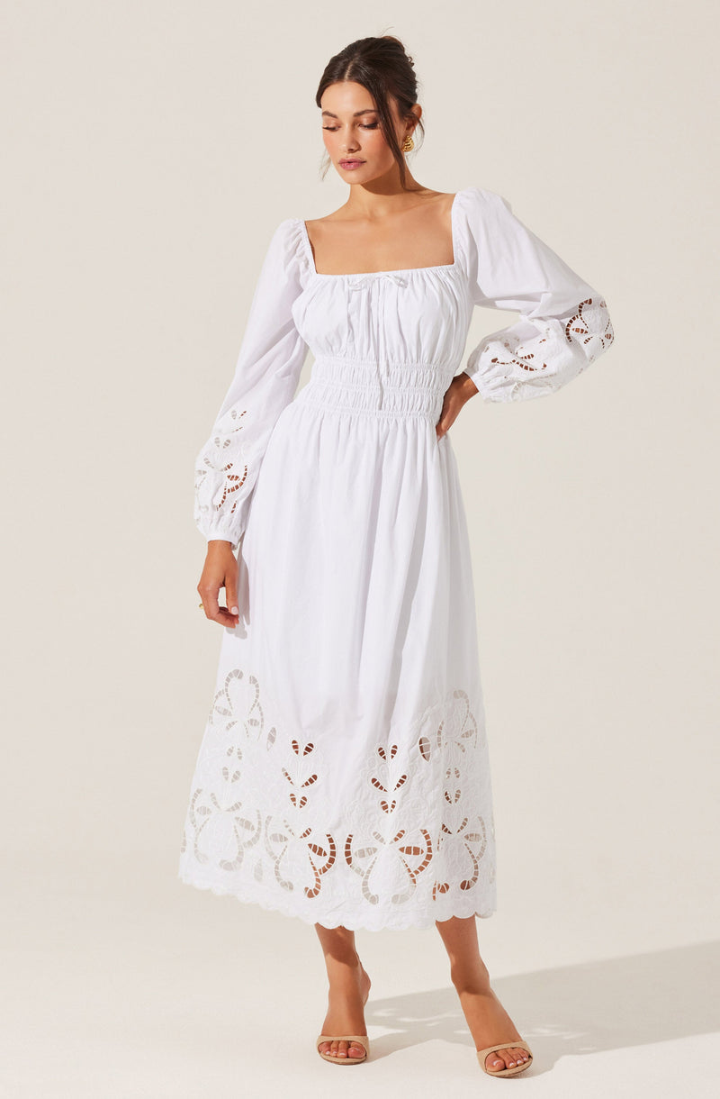 A woman is wearing a long white dress with voluminous, puffed long sleeves that feature intricate cut-out embroidery. The dress has a square neckline and an elasticized waistband that adds shape. The skirt portion is full-length with decorative cut-out embroidery near the hem, creating a delicate and airy look. She is also wearing open-toed heels.