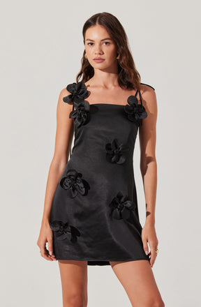 A woman is wearing a black satin mini dress adorned with large 3D floral embellishments. The dress features thin shoulder straps, with some flowers positioned on the straps and others scattered across the front of the dress. The floral decorations are made from the same satin material as the dress, creating a textured, three-dimensional effect.