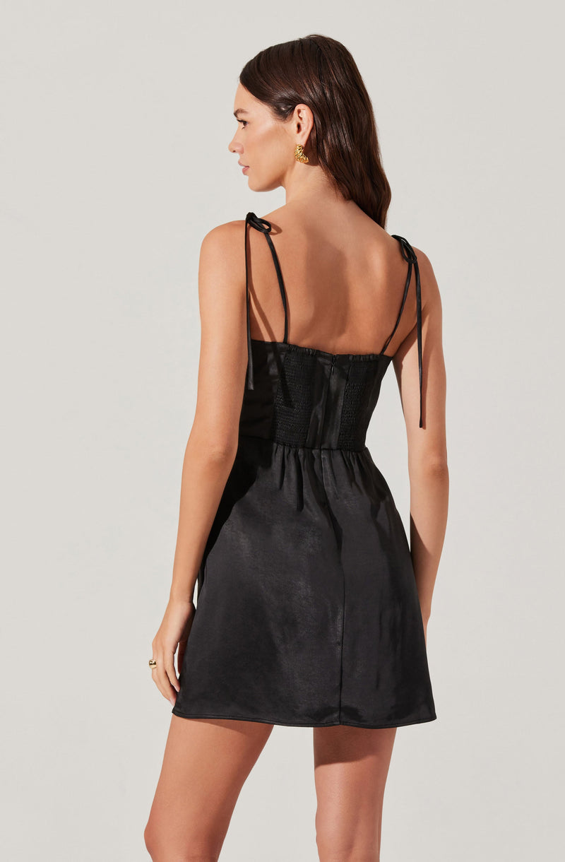 A woman is wearing a black satin dress with spaghetti straps that tie at the shoulders. The dress features a fitted bodice with a sheer panel at the back and a gathered waist, falling to a short hemline above the knees. She has dark brown hair and is accessorizing with gold earrings.