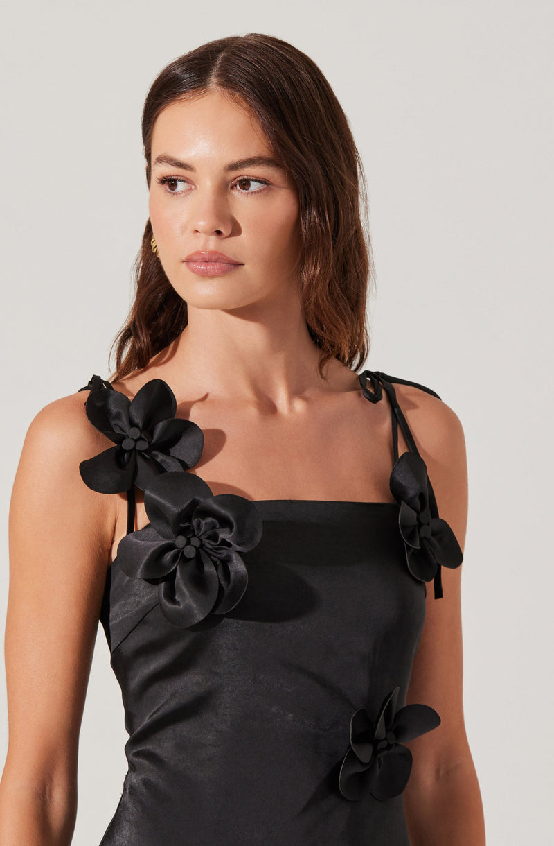 A woman is wearing a black satin dress with thin, adjustable shoulder straps tied into small bows. The dress is decorated with large, black satin floral appliqués on the straps and bodice.