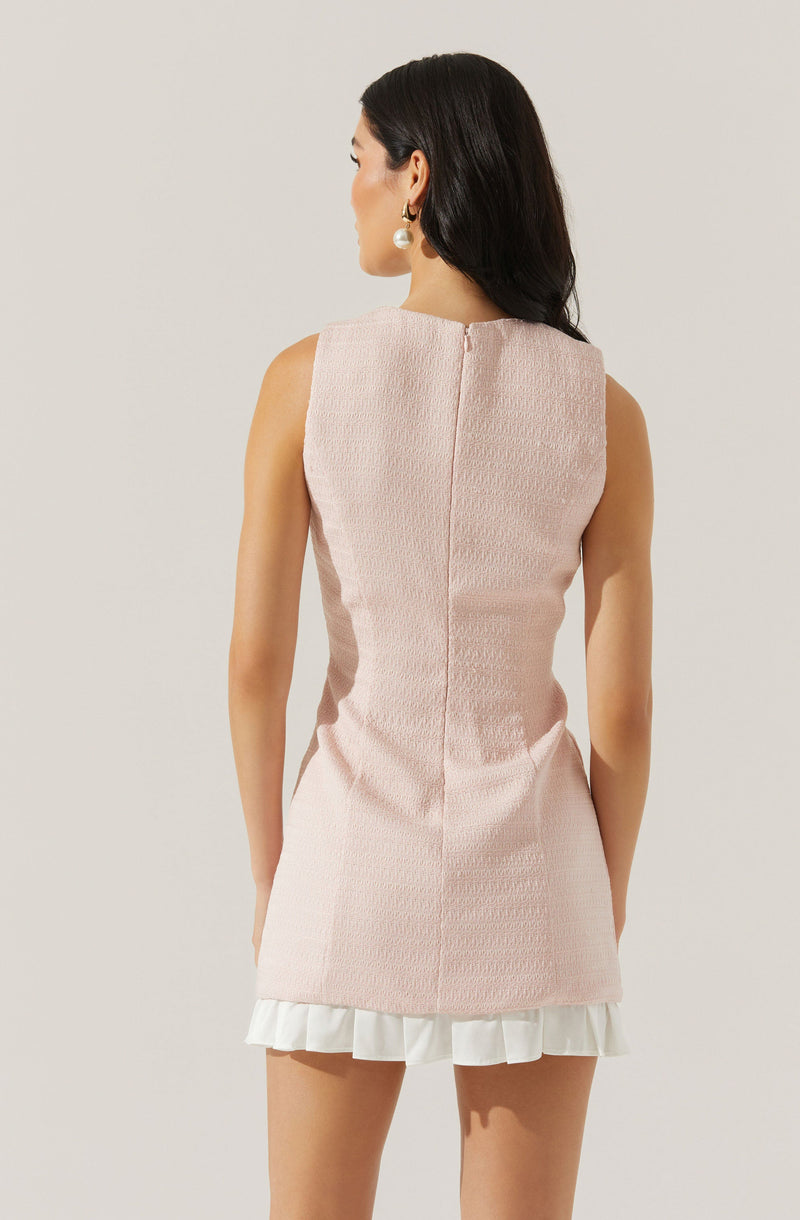 A woman is wearing a sleeveless, light pink textured dress with a straight fit. The dress has a hidden back zipper closure and ends above the knee. The hemline features a white ruffled trim. The woman is accessorized with large pearl drop earrings and has long, dark hair styled in loose waves.