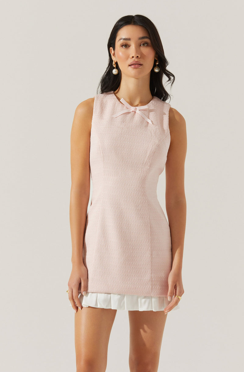 A woman is wearing a sleeveless, light pink textured dress with a small bow at the neckline. The dress is fitted and has a white ruffled hem at the bottom. She is accessorized with large, round earrings and has long, dark hair styled in loose waves.