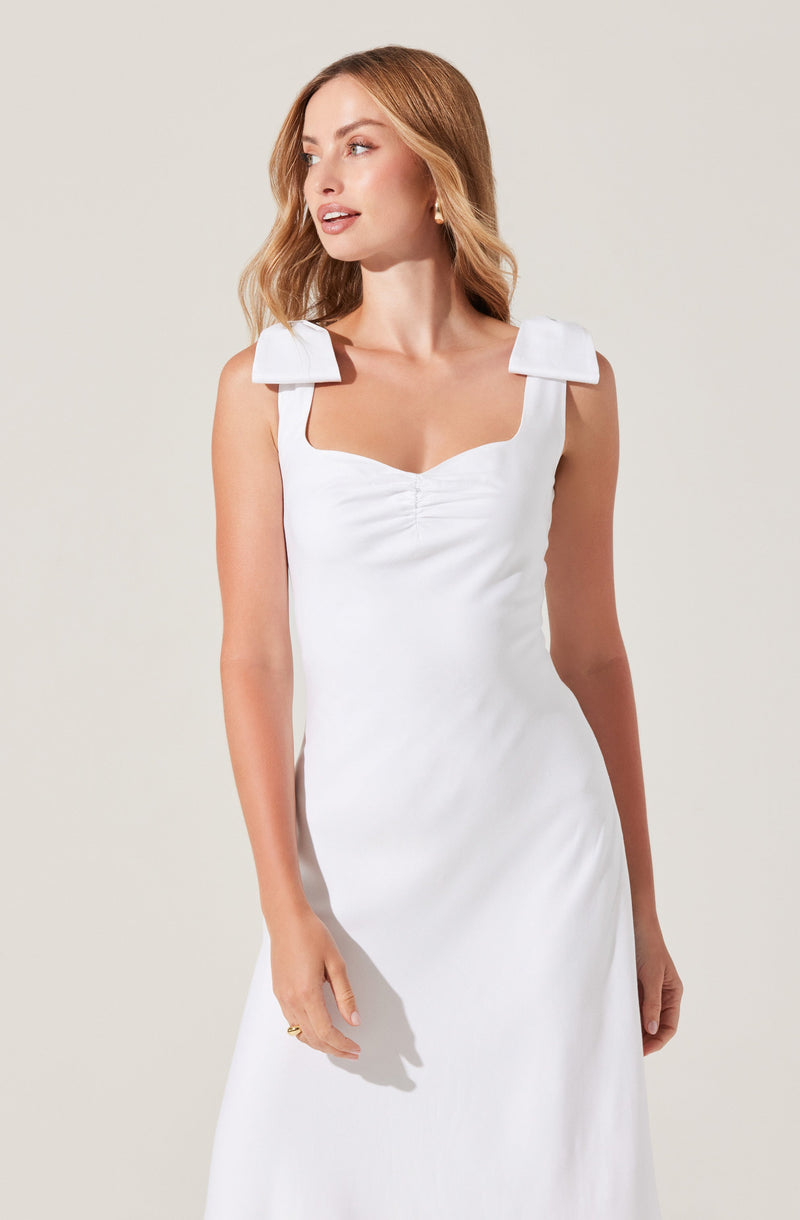 A woman is wearing a sleeveless white dress with wide shoulder straps that have a structured, rectangular design. The dress features a fitted bodice with a subtle ruched detail at the center of the neckline.
