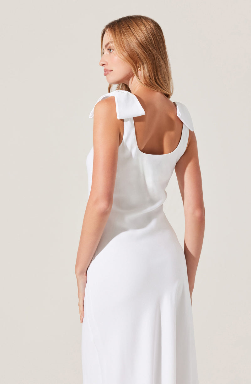 A woman is wearing a white sleeveless dress with large shoulder bows. The dress has a fitted bodice with a rounded neckline and flows down into a long skirt. The fabric appears to be smooth and satin-like.