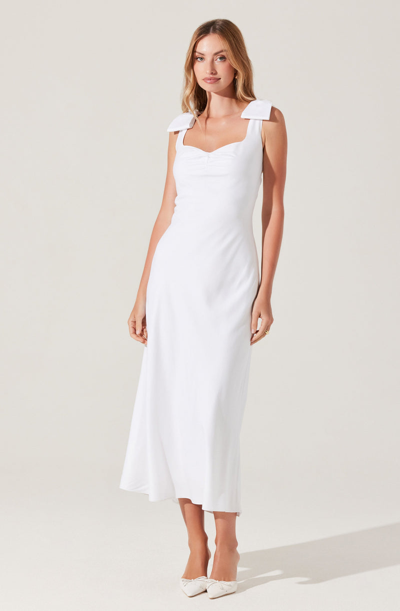 A woman is wearing a white sleeveless dress with wide shoulder straps that feature bow-like details. The dress has a fitted bodice with a sweetheart neckline and falls to a midi length with a straight silhouette. She is wearing white shoes with bows on the front.