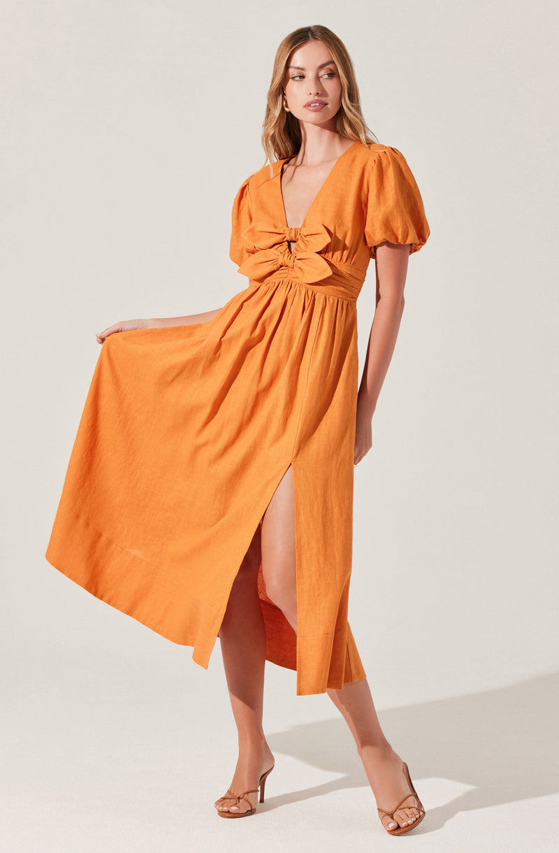 A woman is wearing a bright orange, midi-length dress with short, puffed sleeves and a deep V-neckline. The dress features a gathered detail at the bust with a large bow, and a slit on the side revealing her leg. She is also wearing nude high-heeled sandals.