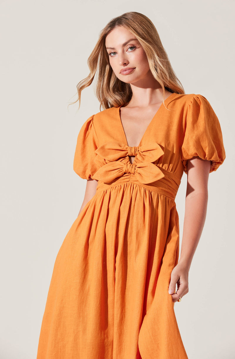 A woman is wearing an orange dress with puffed short sleeves. The dress features a deep V-neckline and a gathered waist with a large decorative bow at the center. The skirt is long and full with gathered fabric flowing down to the floor.