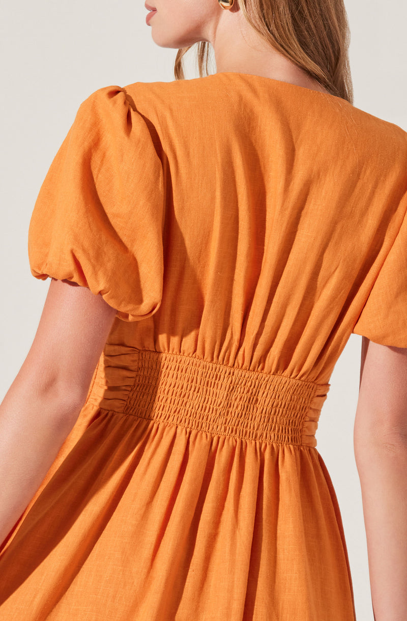 A woman is wearing a burnt orange dress with puffed short sleeves that are gathered at the shoulder. The dress features a fitted, smocked waistband that creates a cinched waistline, and the skirt part of the dress flows out in gathered fabric. The material appears lightweight and slightly textured.