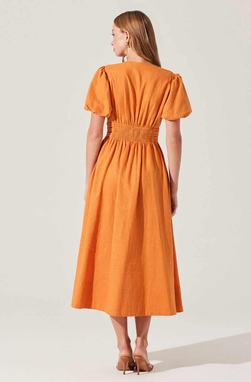 A woman wearing an orange dress with puffed short sleeves, a fitted waist with ruched detailing, and a full-length skirt that reaches the ankles. She is also wearing beige high-heeled shoes.
